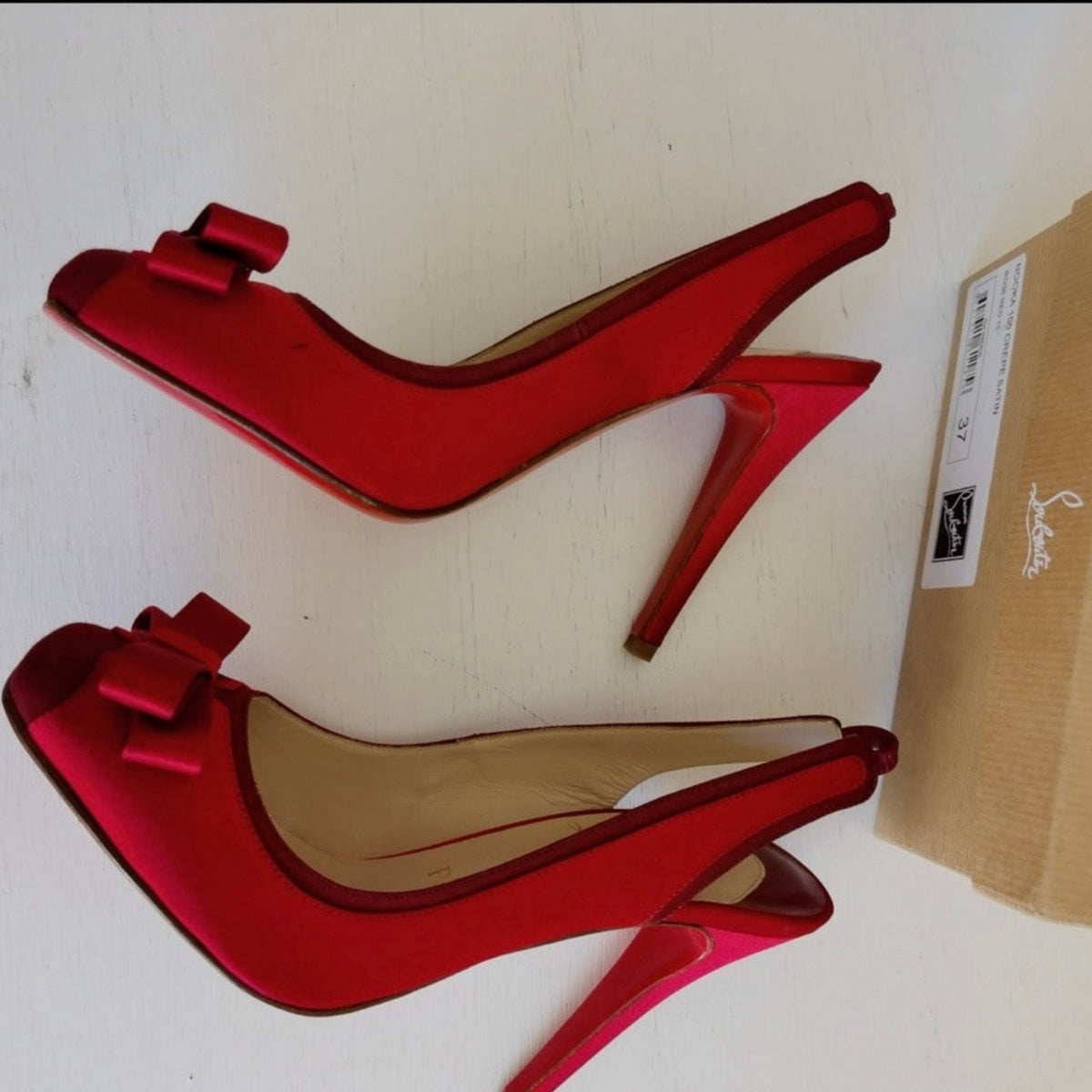 A pair of red satin slingback high heels with bow details on the toes, displayed next to a brown shoebox on a white surface.