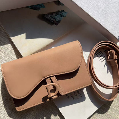 A Dior Saga shoulder bag in a warm tan leather, displayed with its matching strap and branded packaging on a textured beige fabric surface.