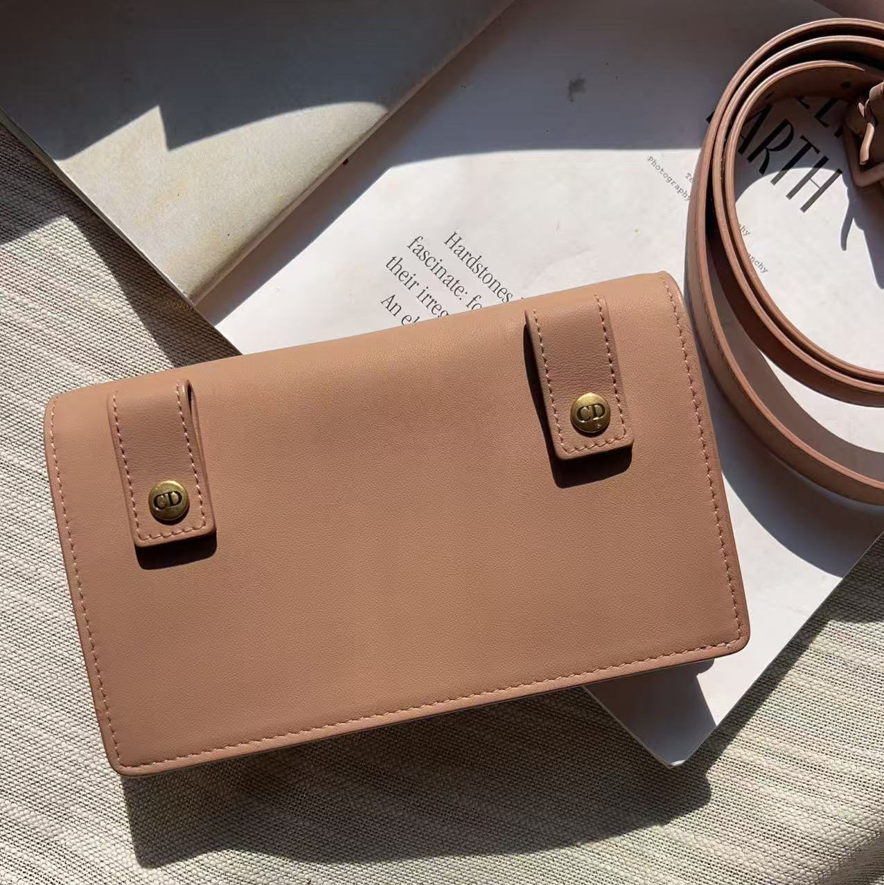 A structured tan leather crossbody bag with gold-tone snap closures and a matching coiled strap, styled on a textured beige surface with a book in the background.