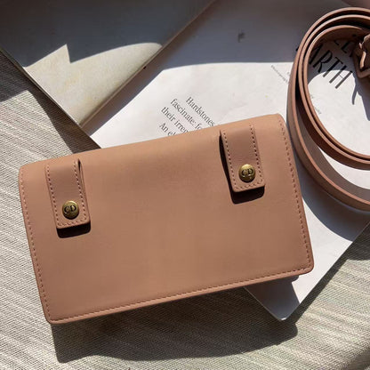 A structured tan leather crossbody bag with gold-tone snap closures and a matching coiled strap, styled on a textured beige surface with a book in the background.