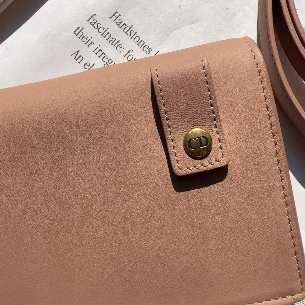 A close-up of a light tan leather wallet or clutch with a flap tab and a gold-tone circular logo button, resting on a white surface with printed paper and a matching strap visible.