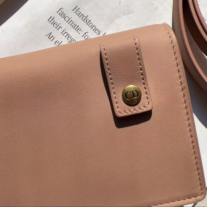 A close-up of a blush pink leather wallet or card holder with a snap-button strap closure, placed on top of a printed sheet. A matching pink belt is partially visible in the upper right, and a neutral surface appears in the upper left.
