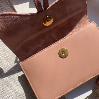 A close-up of a blush pink leather clutch or wallet-style handbag with a matching strap, shown open to reveal a suede-like interior. The exterior has a smooth leather flap with a gold-tone snap button, and the interior has a contrasting brown suede-like lining with a matching gold snap.