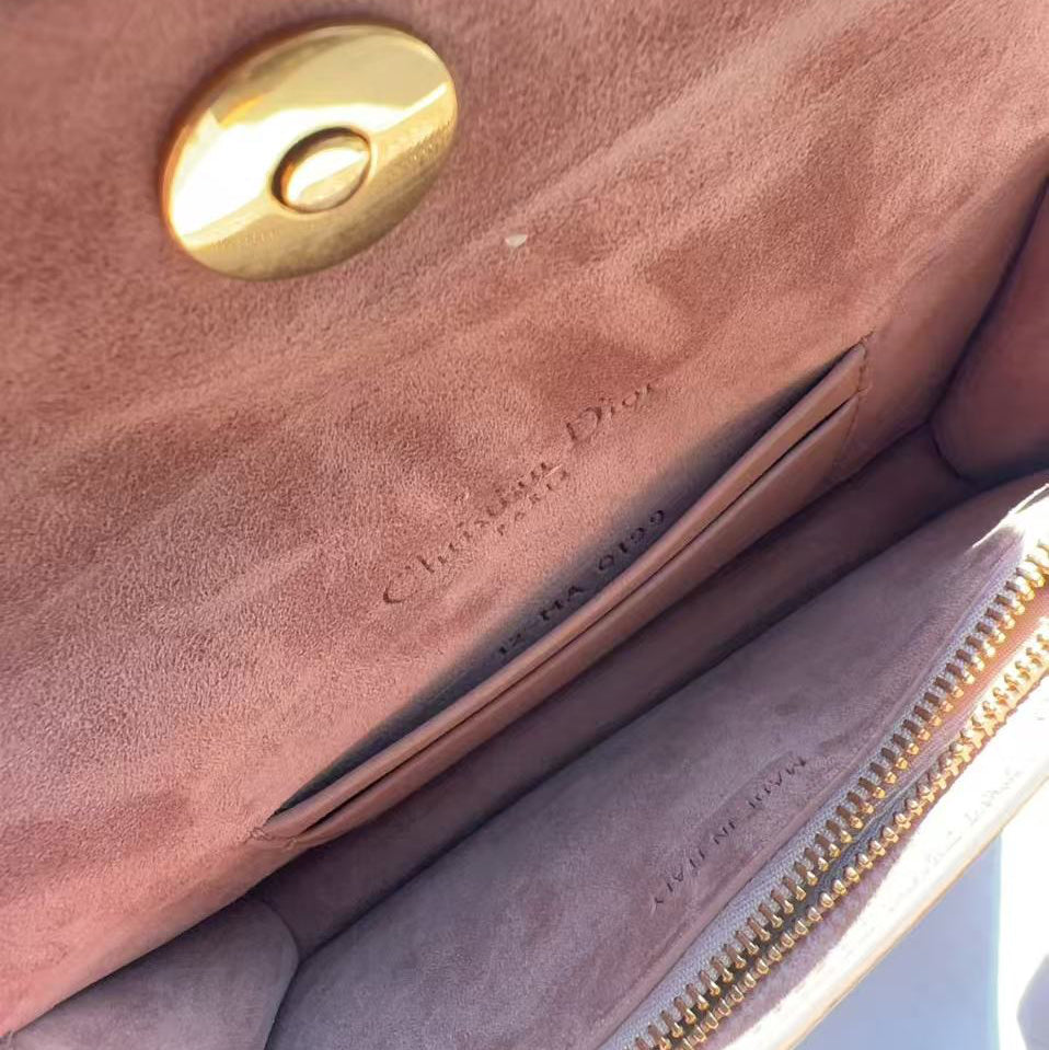 Close-up of the interior of a Christian Dior handbag, showing a dusty rose suede lining, a gold-tone snap button, a zippered pocket, and the embossed Christian Dior name.