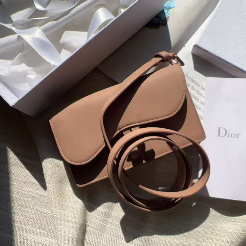 A Dior shoulder bag in a light tan leather, displayed with its white Dior box, dust bag, and brand card on a beige fabric surface.