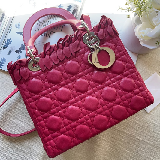 A bright pink quilted leather handbag with a diamond pattern, featuring a ruffled strap detail and gold-tone hardware, laid flat on a light beige fabric surface.