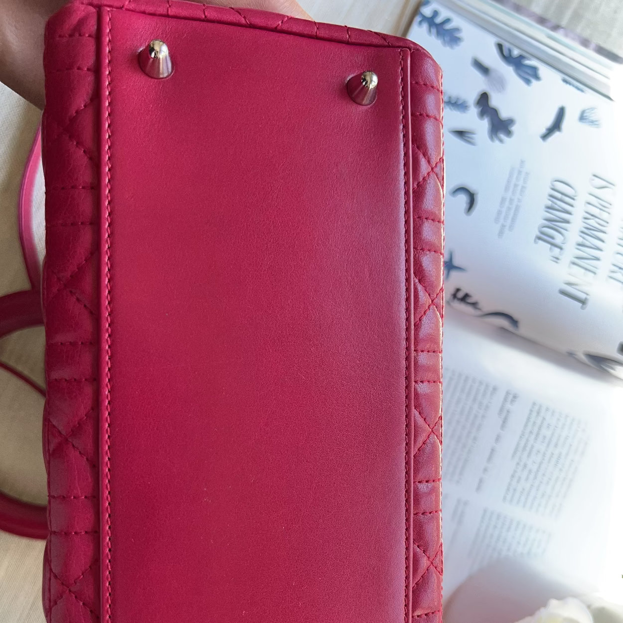 A close-up of a red quilted leather handbag showing the bottom corner with gold-tone metal studs, placed next to an open book and a white rose.