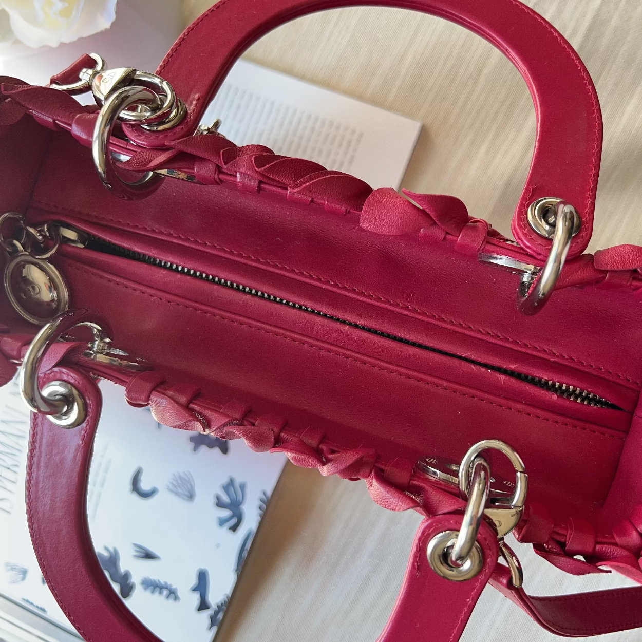 A close-up of a structured red leather handbag with braided trim, gold-tone hardware, and a top handle, photographed on a light fabric surface with a book partially visible underneath.