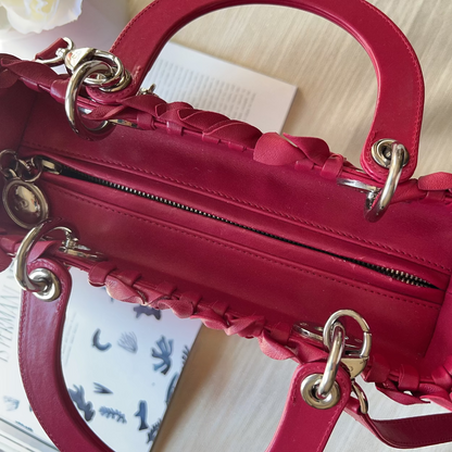 A close-up of a structured red leather handbag with braided trim, gold-tone hardware, and a top handle, photographed on a light fabric surface with a book partially visible underneath.