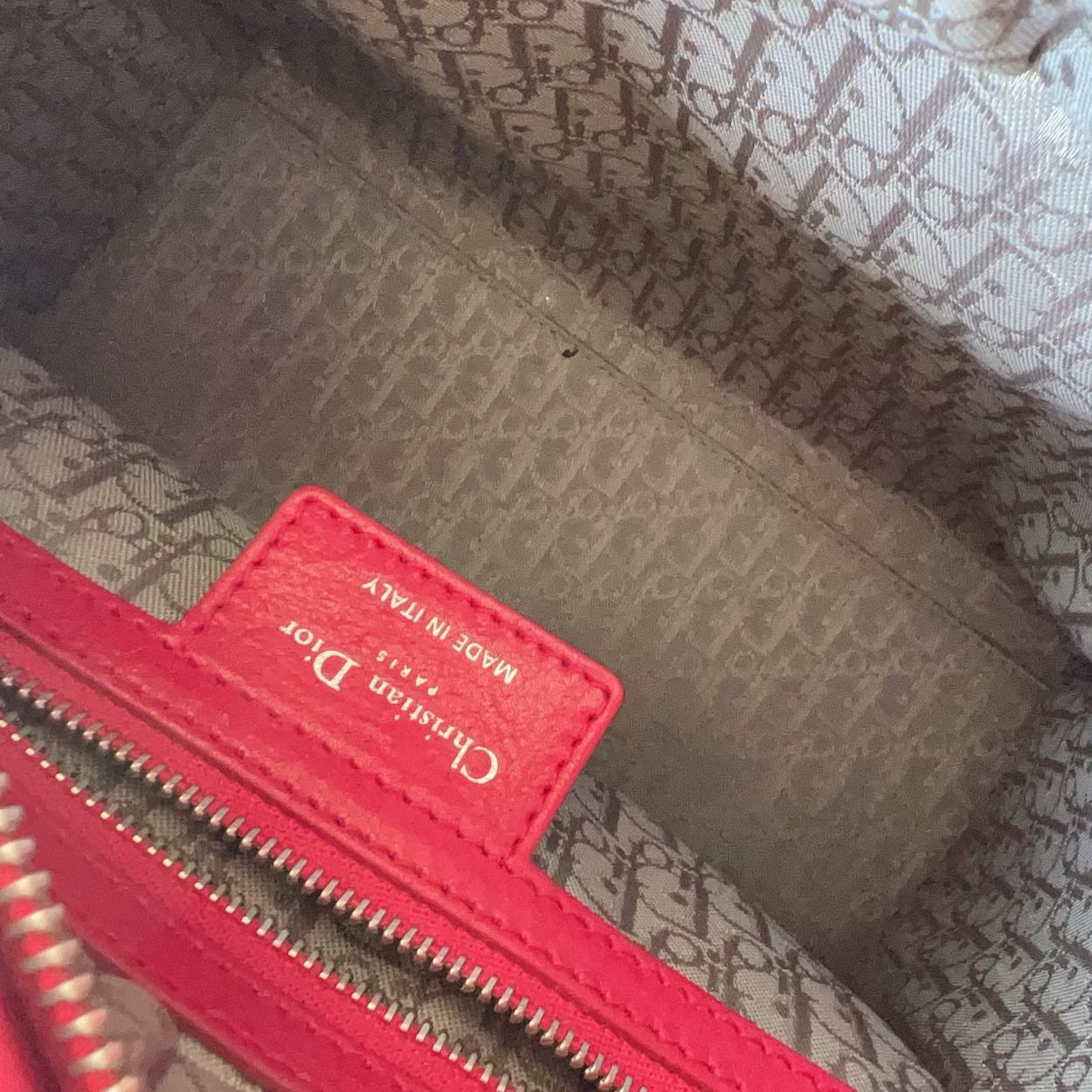 Close-up of the interior of a Christian Dior handbag, showing a red leather interior label with gold lettering, a beige and gray monogram pattern lining, and red zipper trim.