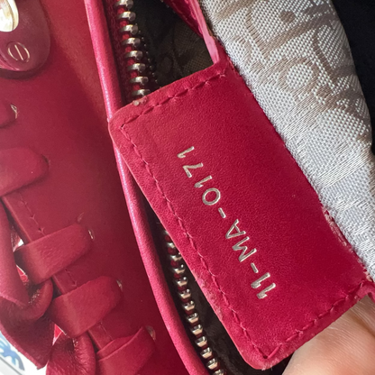 Close-up of a red leather zippered pouch or accessory with a red fabric label reading '11-MA-0171' in gold lettering, attached to a red laced item. A patterned gray fabric and a finger are also visible.