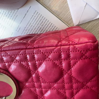 A close-up of a quilted fuchsia pink leather handbag with a circular gold-tone hardware detail, resting on a beige textured surface next to an open book and a white envelope.