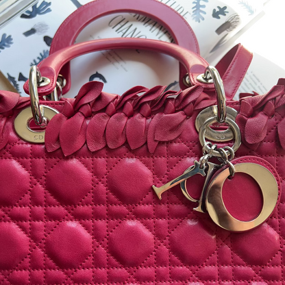 A close-up of a bright pink quilted leather handbag with a ruffled top border, gold-tone hardware, and a hanging charm, photographed on a light surface with printed papers in the background.