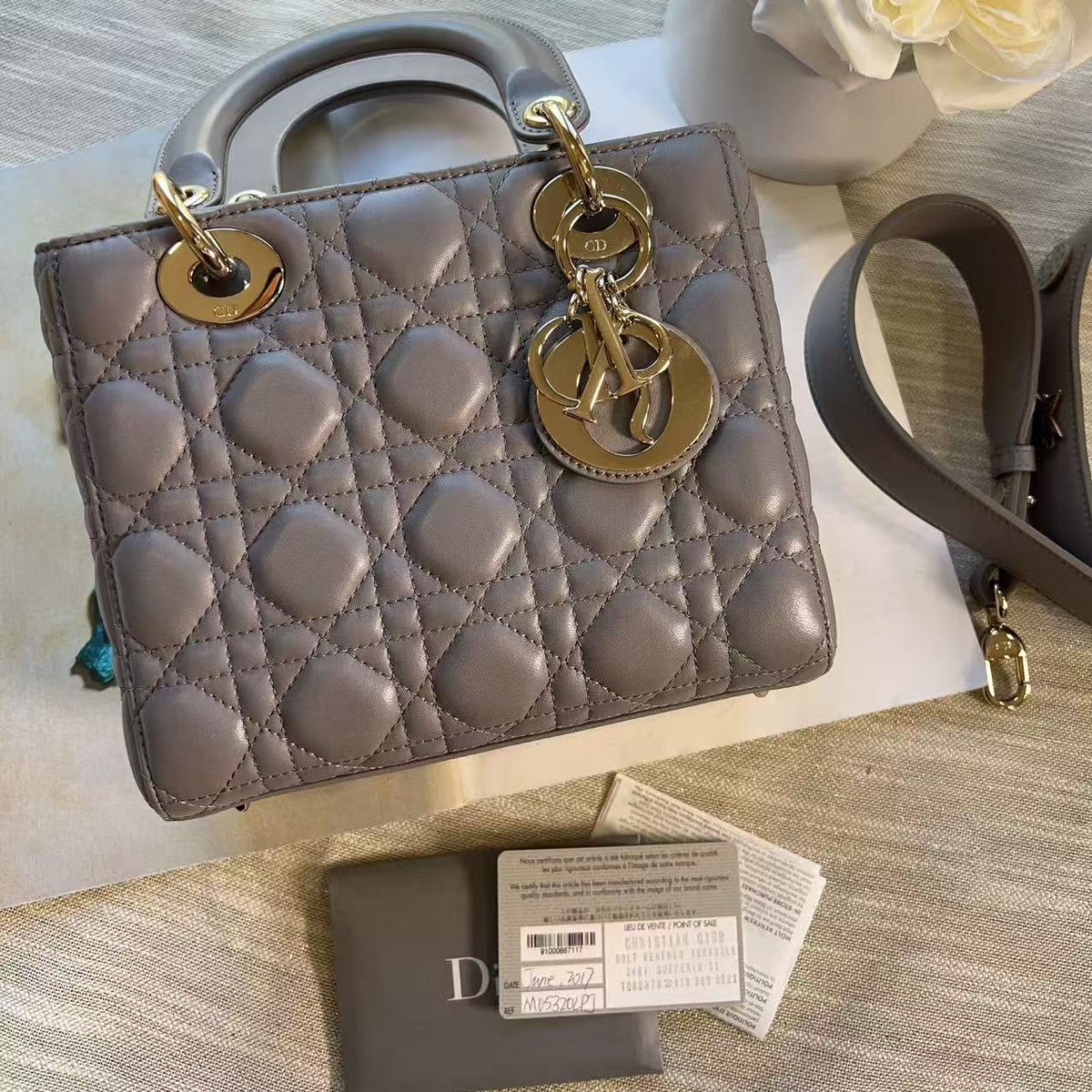 A quilted taupe-gray Christian Dior Lady Dior handbag with gold-tone hardware, displayed on a light-colored surface with its dust bag and tags in front, and white flowers in the background.