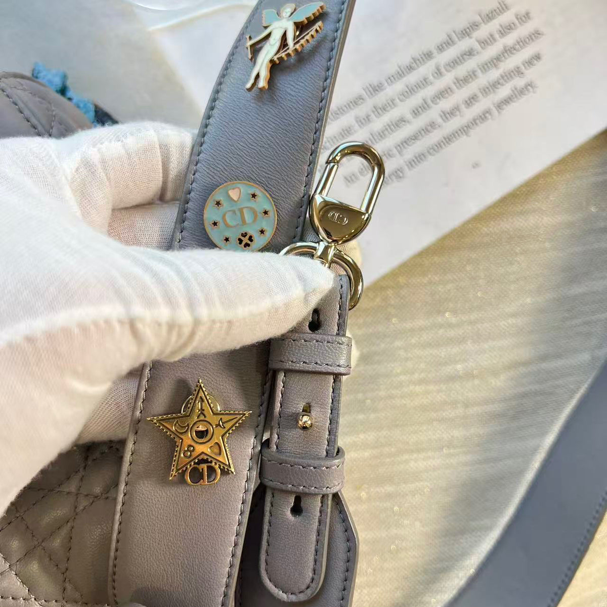 A close-up of a light gray quilted handbag strap with decorative gold-tone charms, including a star-shaped pendant, a round medallion, and a butterfly charm, held by a white-gloved hand.