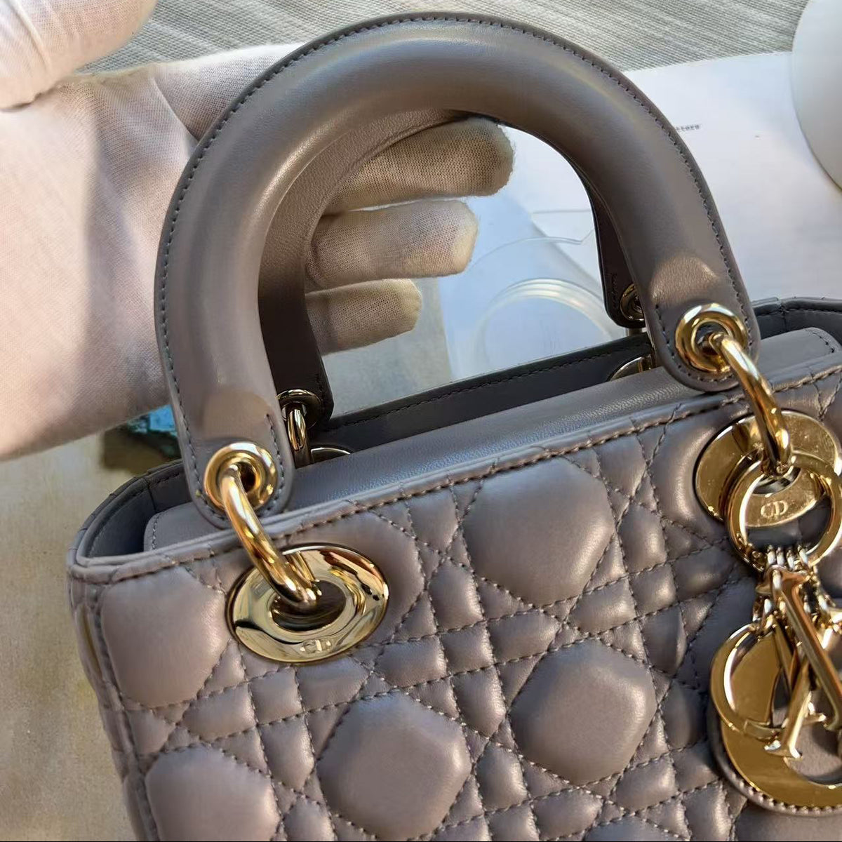 A close-up of a light gray quilted leather handbag with gold-tone hardware and a curved top handle, held by a gloved hand against a light gray fabric background.