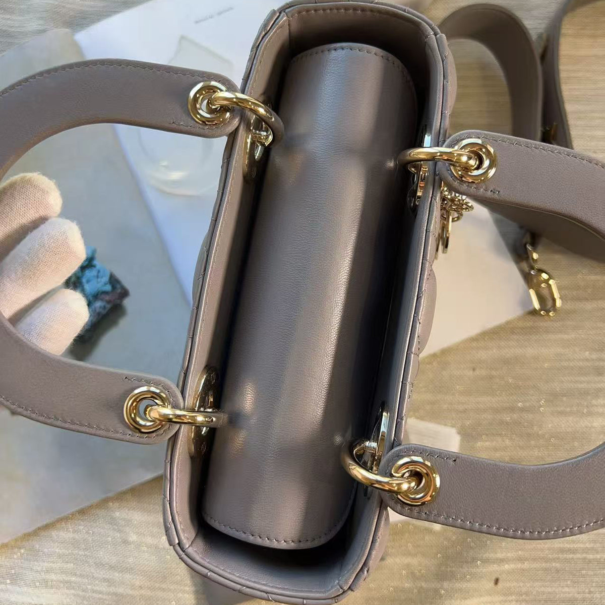 A top-down view of a structured handbag in a muted taupe-gray color with gold-tone hardware, shown on a light wood surface with a white flower vase in the background.