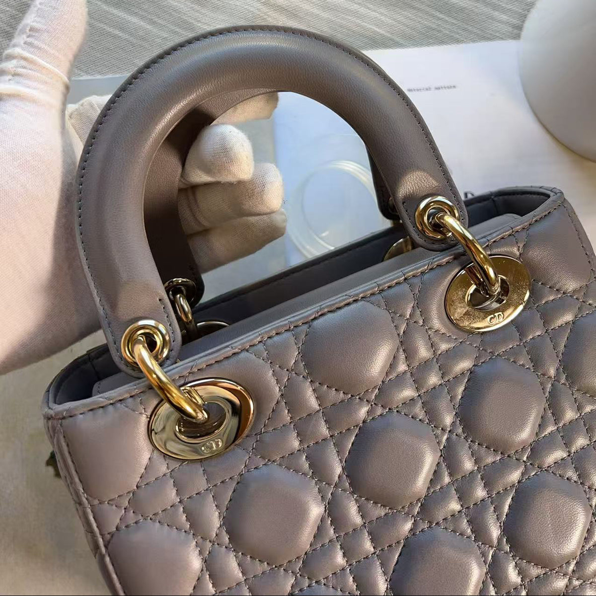 A close-up of a structured quilted handbag in a muted taupe-gray color with gold-tone hardware and a curved top handle, held by a gloved hand on a light surface.