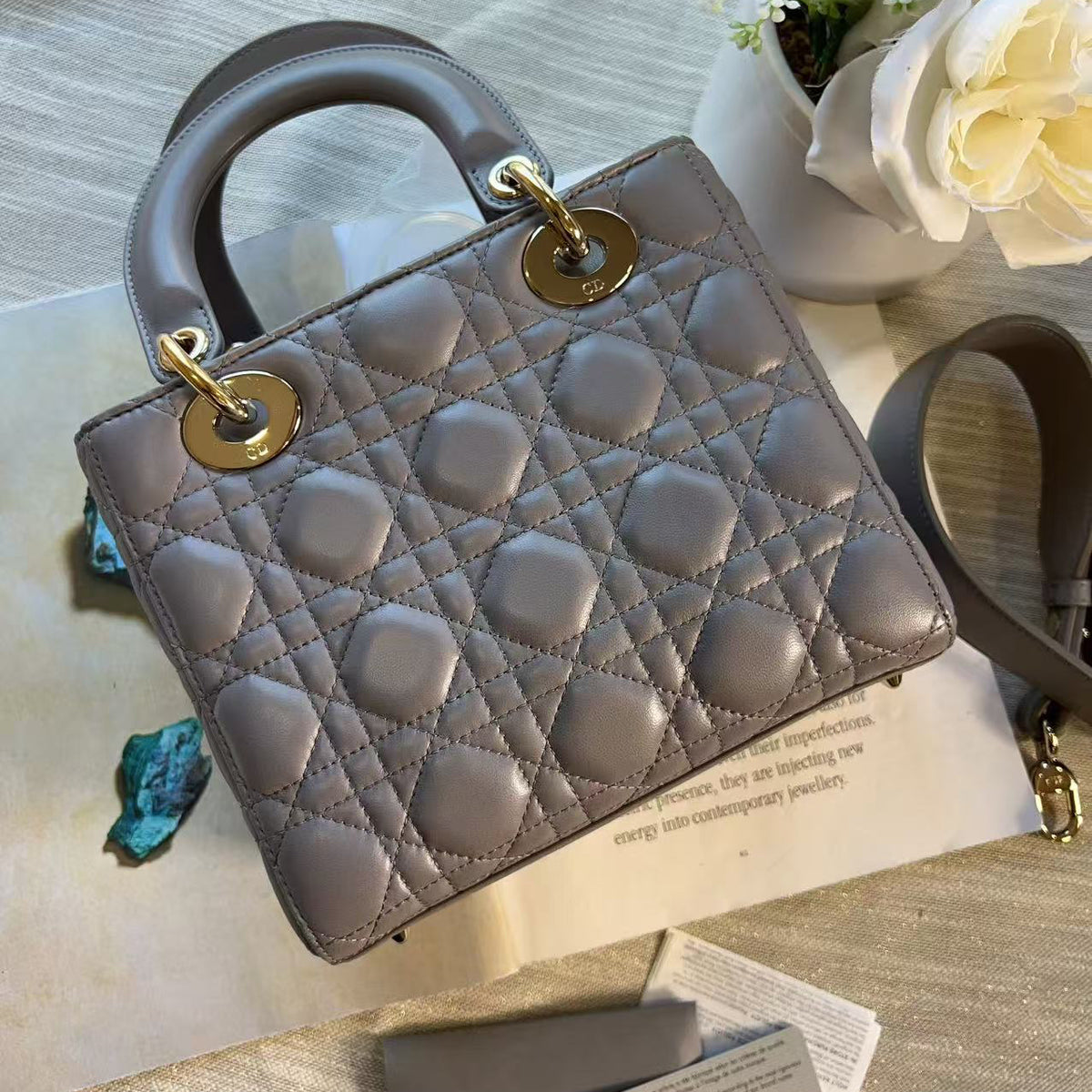 A structured Christian Dior Lady Dior handbag in taupe gray patent leather with gold-tone hardware, displayed on a light wood surface with a magazine, dust bag, and white roses in the background.