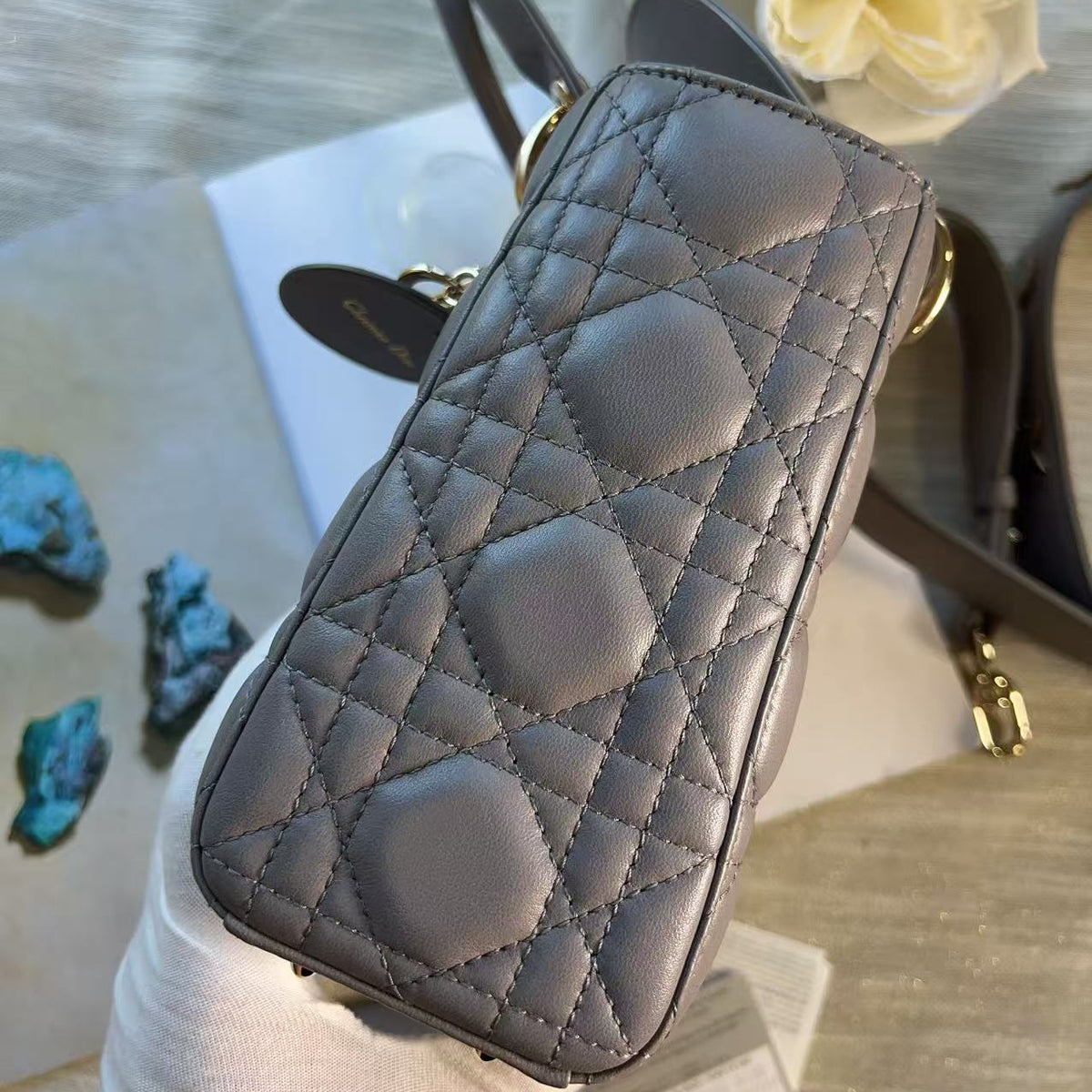 A close-up of a quilted gray leather handbag held by a gloved hand, showing the diamond-stitched pattern and gold-tone hardware, with flowers and a neutral tabletop in the background.