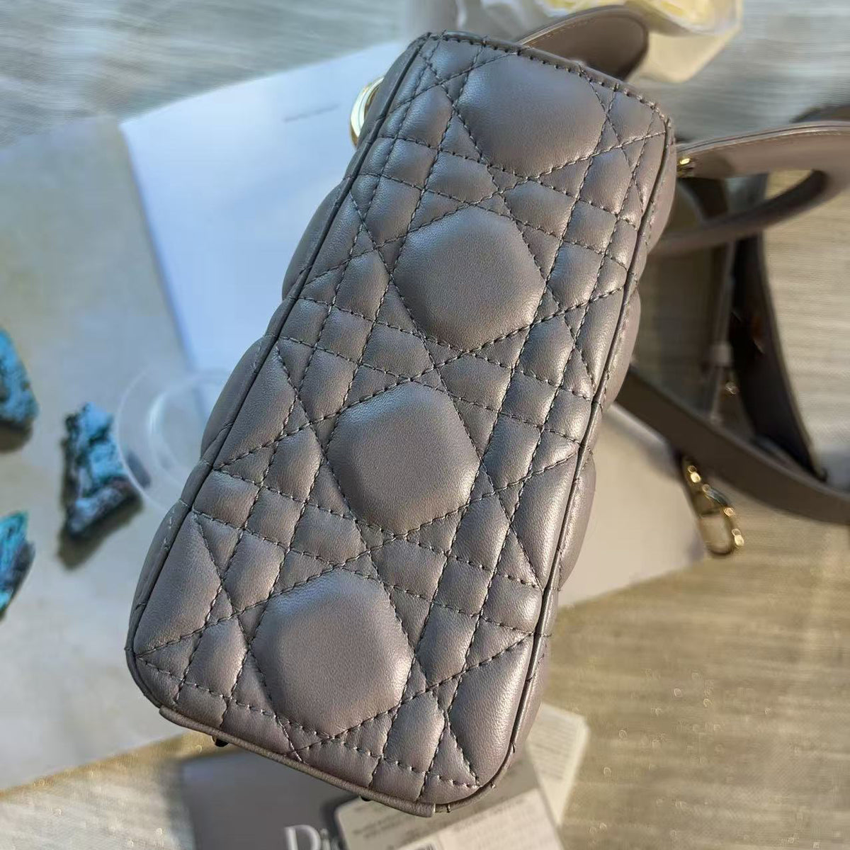 A close-up of a quilted gray leather pouch or wallet with a diamond pattern, resting on a light wood surface with a Dior card and white flowers in the background.