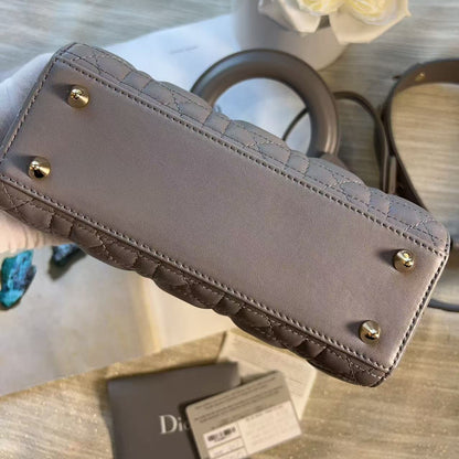 A close-up of a taupe quilted handbag showing the bottom corner with gold-tone studs, held by a hand over a light wood surface with a Dior card and white flowers in the background.