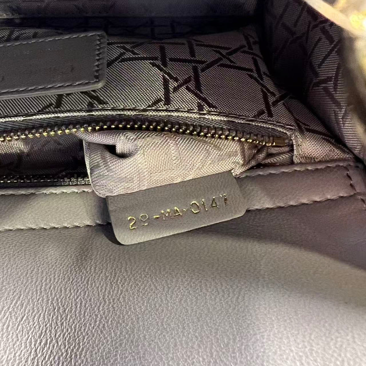 Close-up of a gray textured leather bag interior showing a zipper, patterned lining, and a small leather tag with gold-tone lettering.