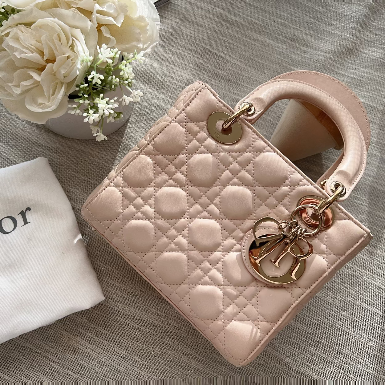 A pale blush pink quilted leather Lady Dior handbag with gold-tone hardware, styled on a textured gray fabric alongside a white Dior dust bag and cream flowers.
