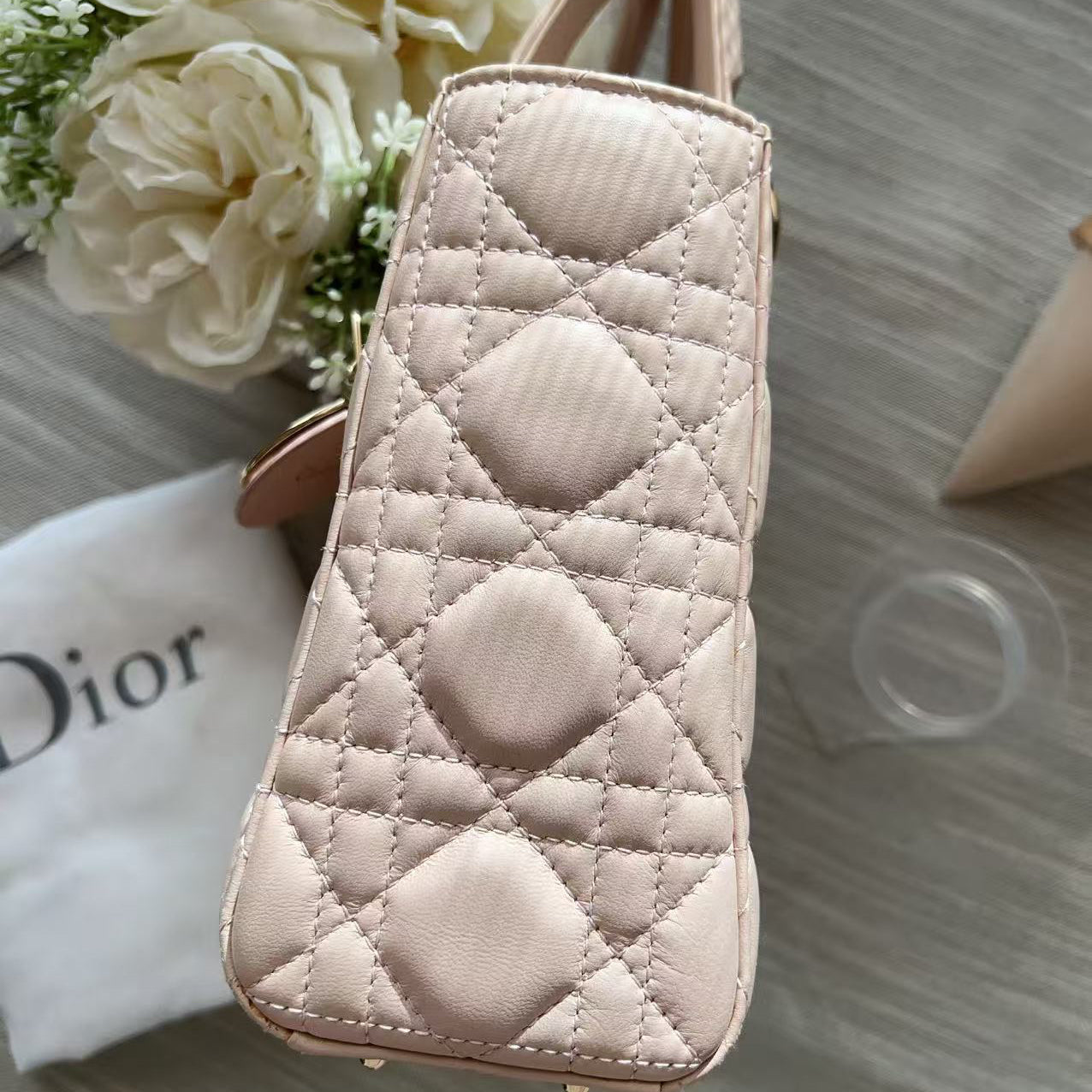 A light pink quilted handbag with diamond stitching, shown on a gray wood-textured surface next to a white Dior dust bag and white flowers.