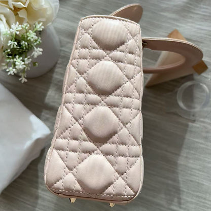 A light pink quilted leather handbag with a diamond pattern and gold-tone hardware, shown from a top-down angle on a gray wood-textured surface with decorative props.