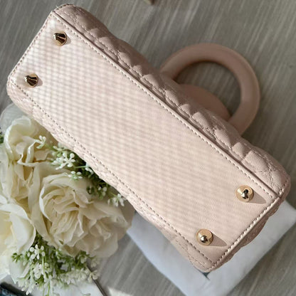 A light beige quilted leather handbag with gold-tone studs and a circular handle, styled on a light wood surface next to white flowers.