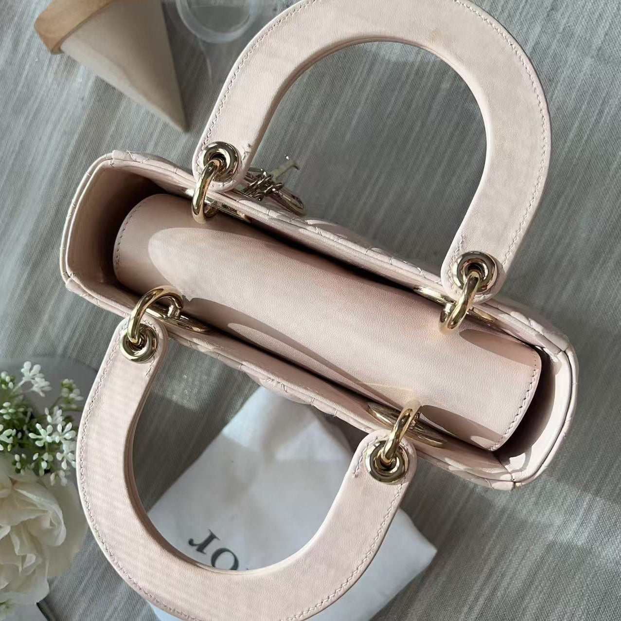 A light pink leather handbag with gold-tone hardware, shown from an overhead angle on a gray textured surface with a white flower and a white dust bag nearby.