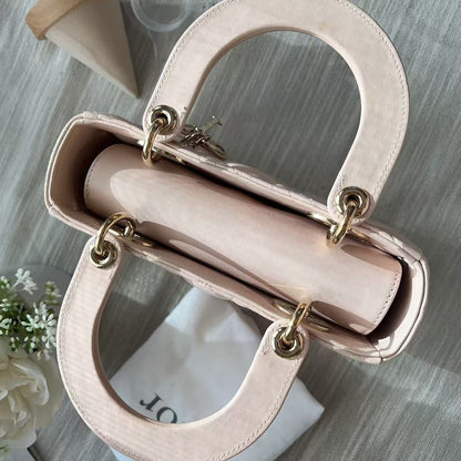 A light pink leather handbag with gold-tone hardware, shown from an overhead angle on a gray textured surface with a white flower and a white dust bag nearby.