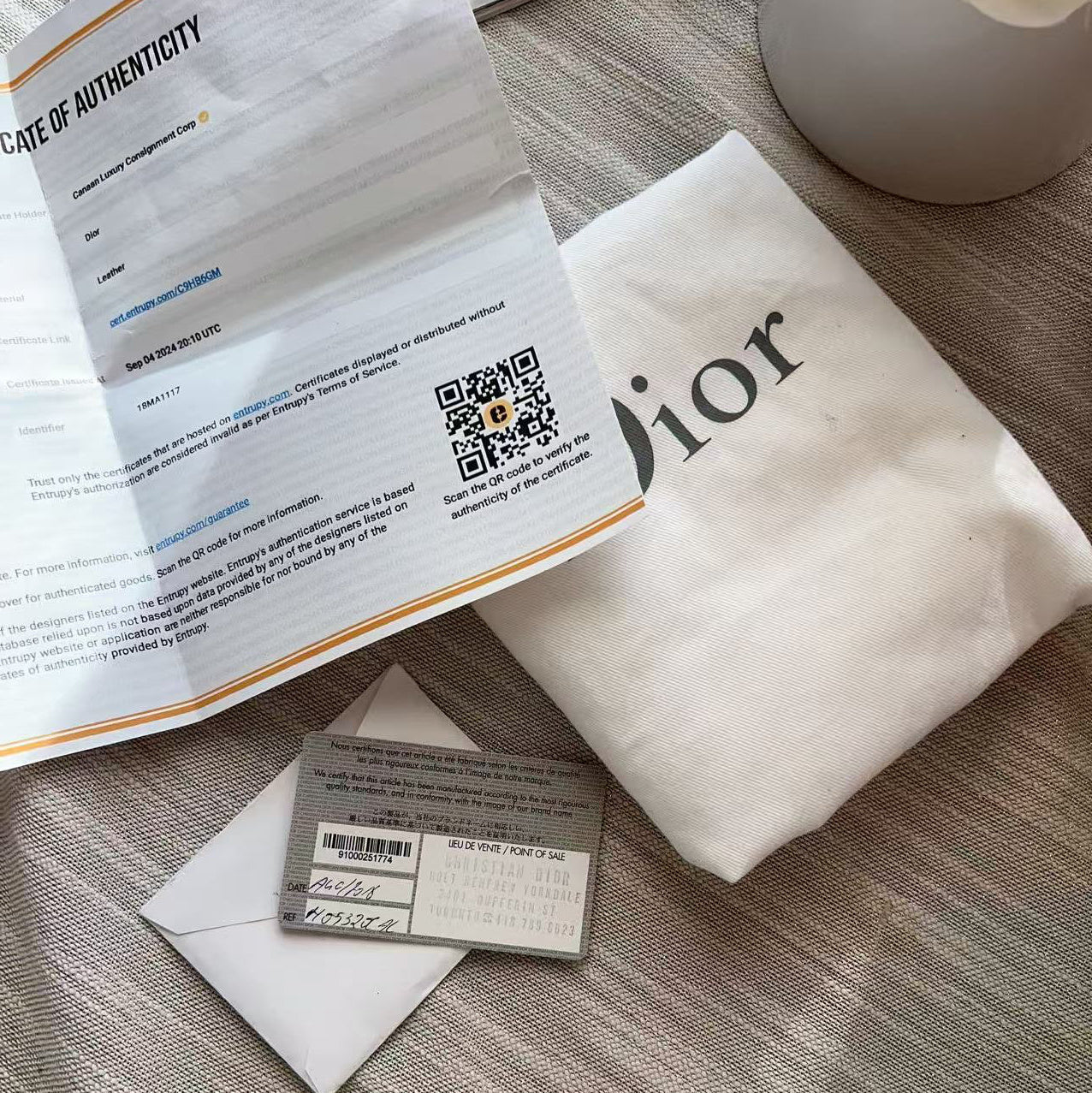A Dior-branded cream-colored dust bag lies on a textured beige fabric surface next to an open printed document, a small gray ticket or card, and a white envelope.