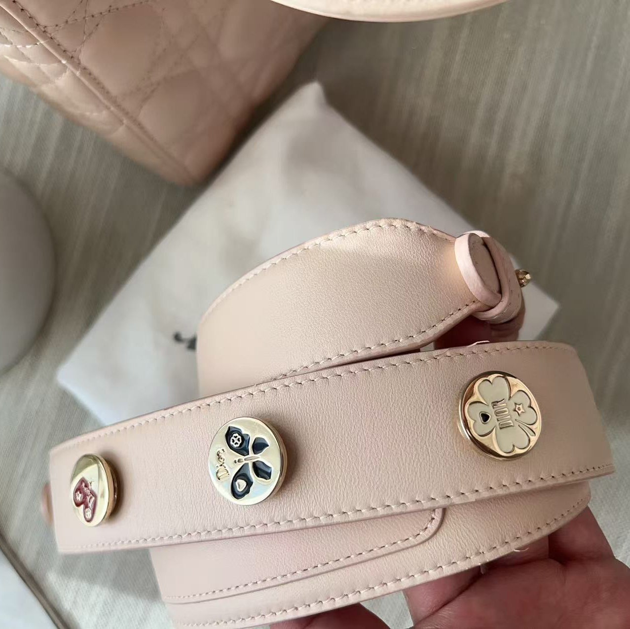 A close-up of a light blush pink leather shoulder strap with decorative round metal studs, held by a hand over a white dust bag with a quilted bag and flowers in the background.