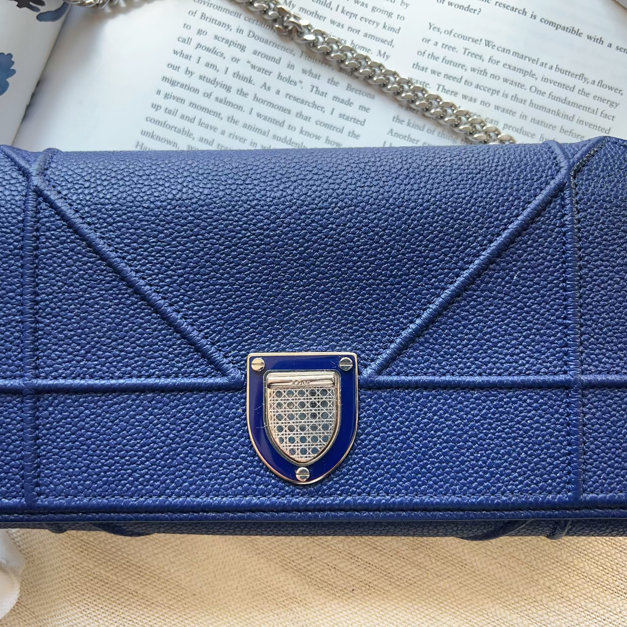 A blue pebbled leather clutch purse with a geometric quilted design and a silver-tone metal clasp, resting on an open book with a chain strap visible.