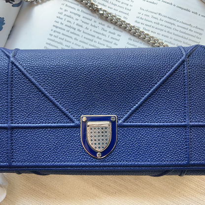 A blue pebbled leather clutch purse with a geometric quilted design and a silver-tone metal clasp, resting on an open book with a chain strap visible.
