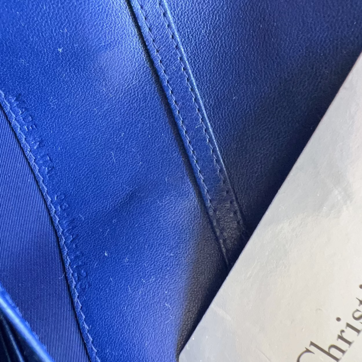 Close-up of a royal blue leather bag with visible stitching and a white paper tag printed with 'Christian Dior'.