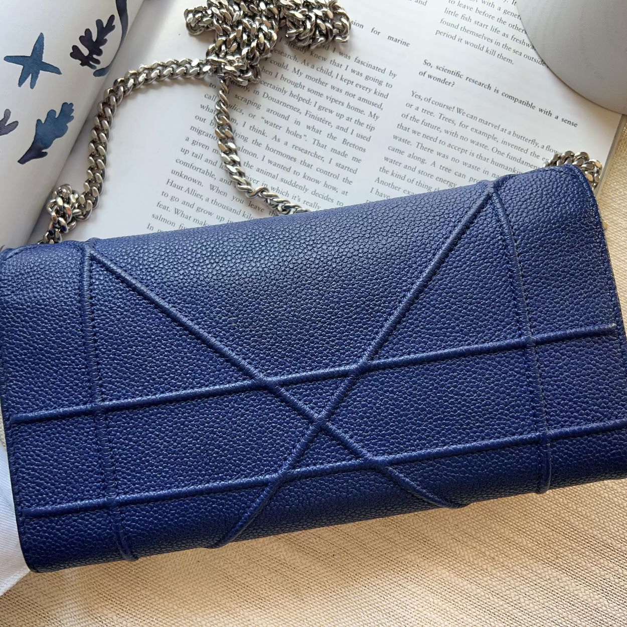 A navy blue textured leather clutch bag with a geometric quilted pattern and a silver chain strap, styled on a light wood surface with an open book and a white fabric accessory.