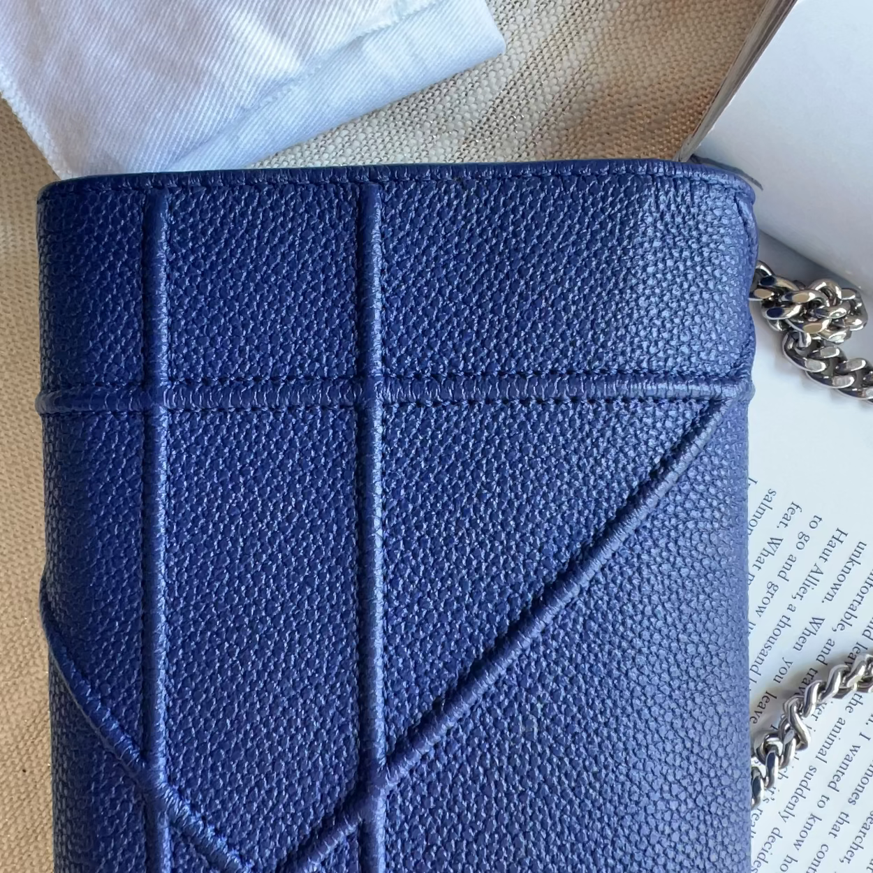 A close-up of a blue pebbled leather clutch or wallet-style handbag with a quilted geometric pattern and a silver chain strap, styled on a beige fabric surface with a book and white fabric in the background.