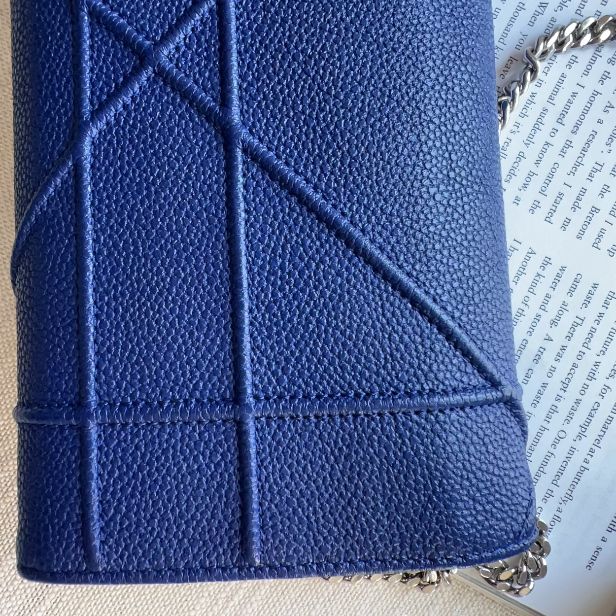 A close-up of a blue pebbled leather handbag with diagonal quilted stitching, resting on a textured beige fabric surface next to an open book.