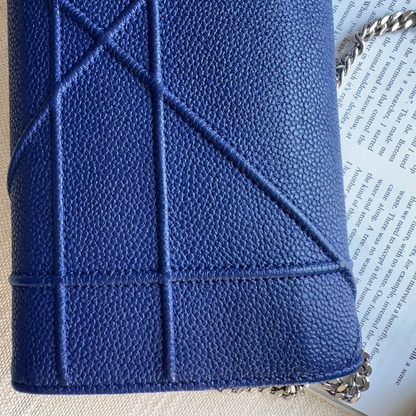 A close-up of a blue pebbled leather handbag with diagonal quilted stitching, resting on a textured beige fabric surface next to an open book.