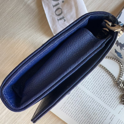 A close-up of a blue pebbled leather wallet or clutch shown from the inside, with a gold-tone clasp and silver chain visible, resting on a cream fabric surface next to printed papers.