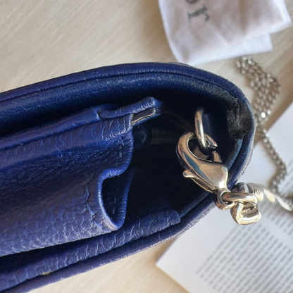 Close-up of a blue leather handbag showing the interior and a silver-tone lobster clasp, with part of a chain strap and a light wood surface in the background.