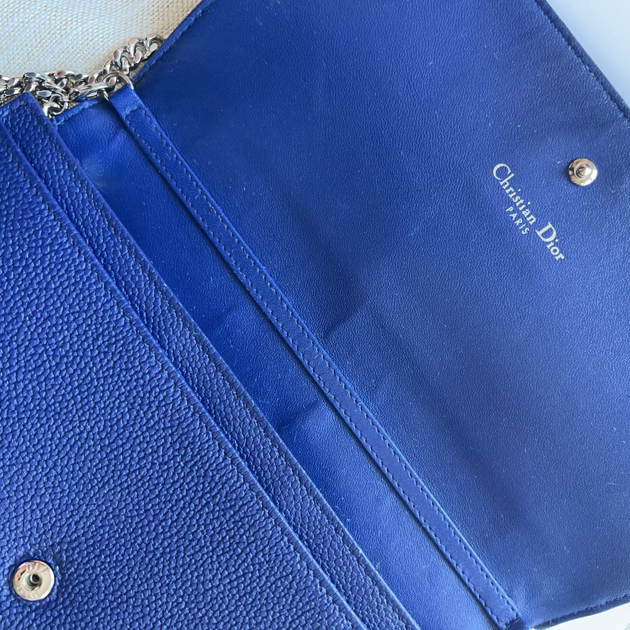 A close-up of a royal blue Christian Dior clutch or envelope-style handbag, showing textured leather on the left, smooth satin trim, a chain strap, and the white Dior logo printed inside the flap.