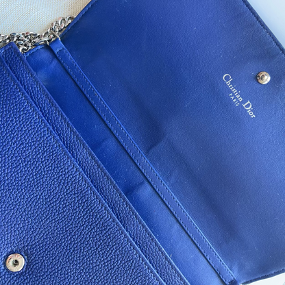 A close-up of a royal blue Christian Dior clutch or envelope-style handbag, showing textured leather on the left, smooth satin trim, a chain strap, and the white Dior logo printed inside the flap.