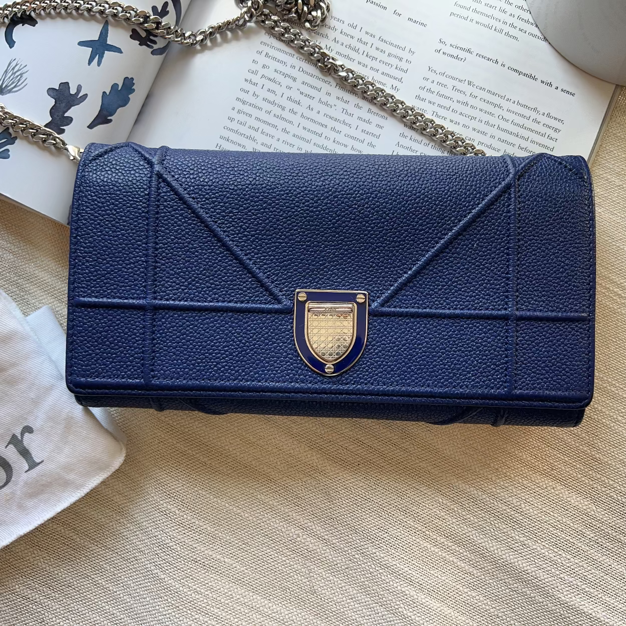 A navy blue textured leather clutch bag with a gold-tone dome clasp, styled on a light wood surface with an open book, a white Dior-branded dust bag, and a chain strap.