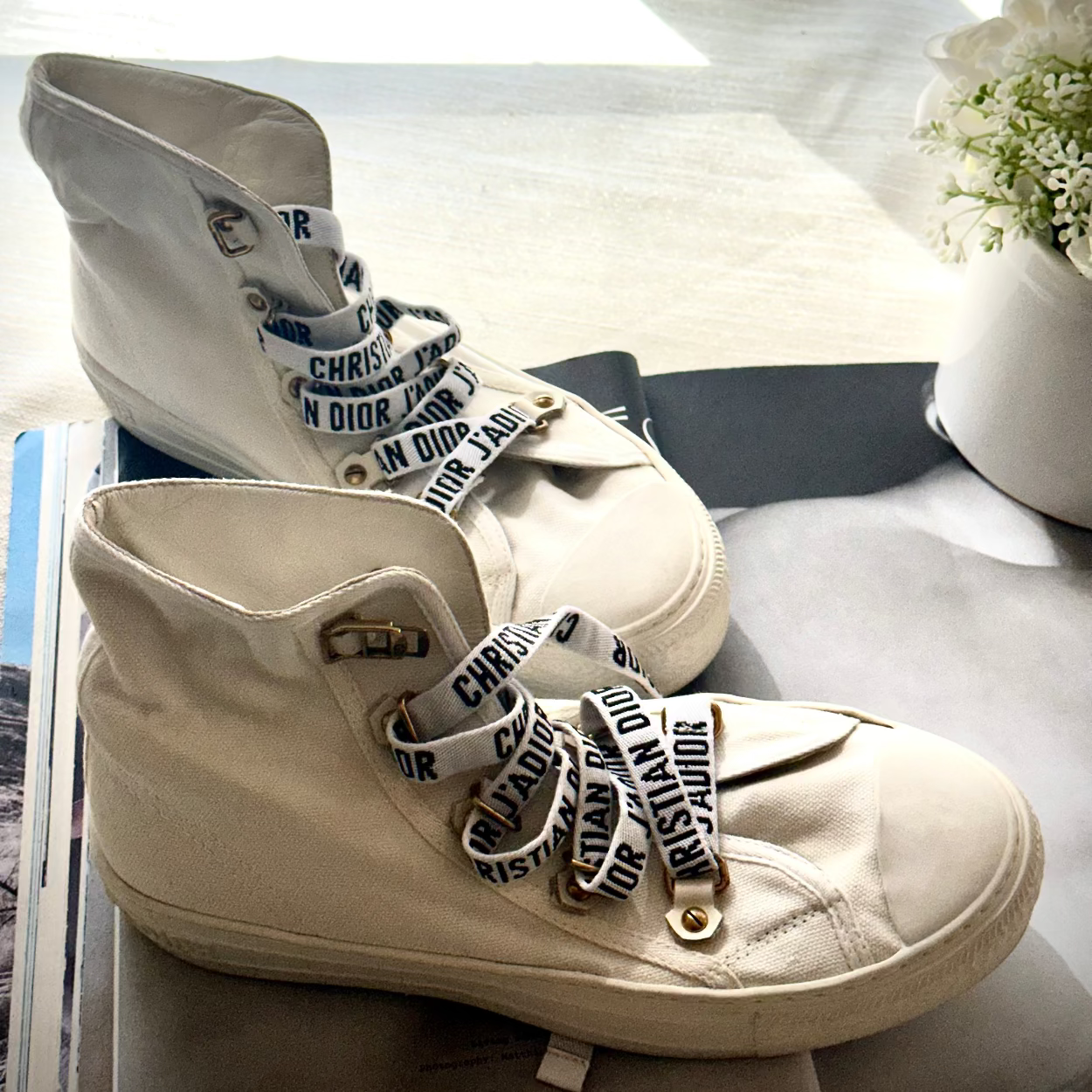 A pair of off-white high-top canvas sneakers with black-and-white Dior text ribbon laces, displayed on a tabletop near a window.