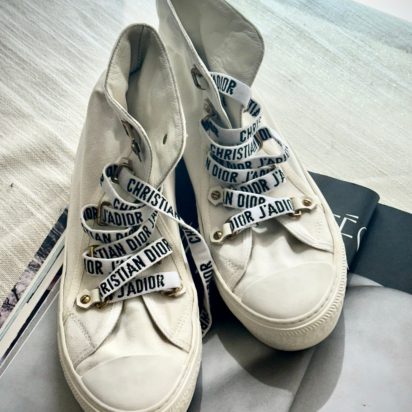 A pair of off-white high-top canvas sneakers with wide white laces printed with 'CHRISTIAN DIOR' in black lettering, resting on a gray surface with a black Dior box.