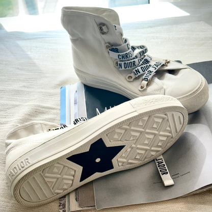 A pair of white high-top Christian Dior sneakers with navy blue star details on the soles and white laces printed with the Dior name, displayed on a beige fabric surface with papers underneath.